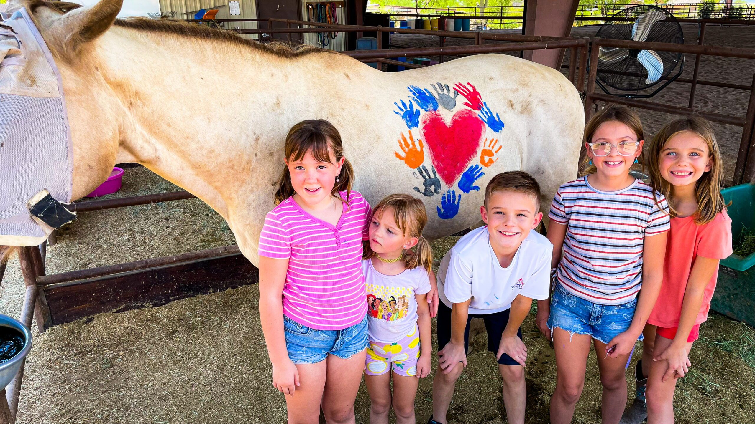 A group of children standing in front of a horse that has different colored hand prints surrounding a heart painted on its side.