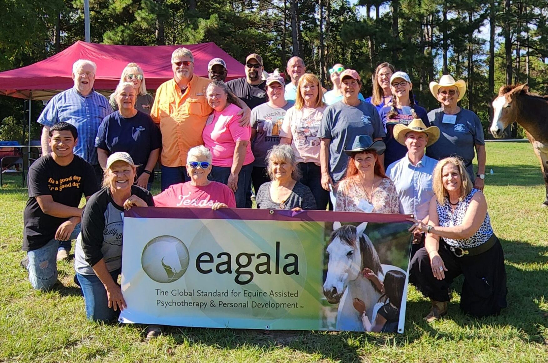 A group of Eagala therapists and clients posing with the Eagala banner with horses and trees in the background